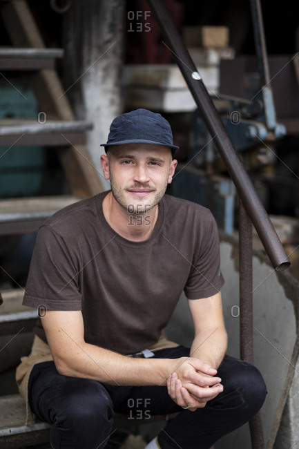 Male barista in black baseball cap and face mask sitting on stairs outdoors, smiling at camera.