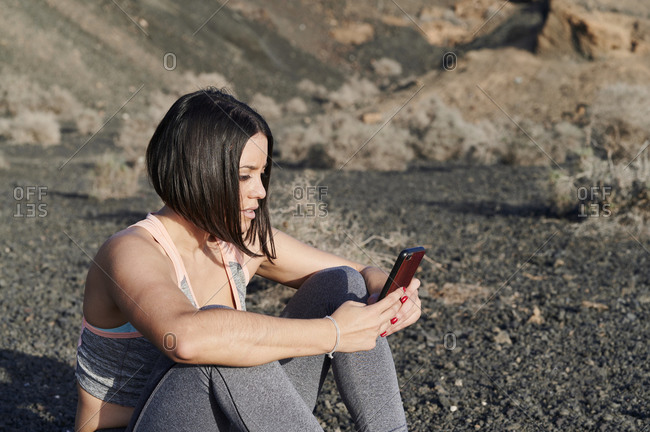 Fit young woman in sportswear sitting outside in some rocky terrain and checking her phone before a workout session