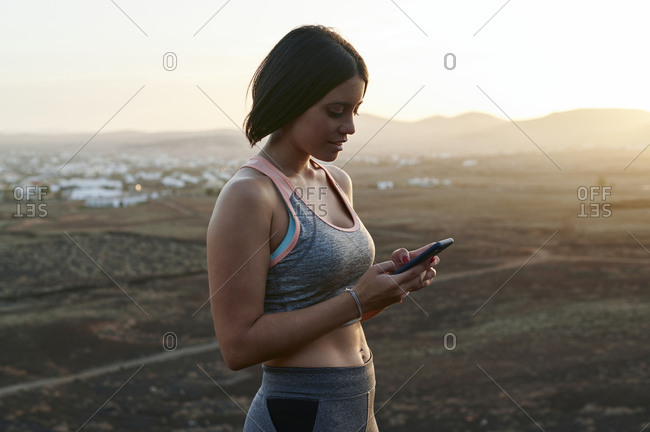 Fit young woman in sportswear standing outside in the countryside at sunset and checking her phone after a workout