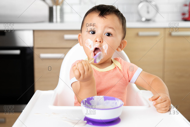 Cute messy baby in high chair eating yogurt with funny face