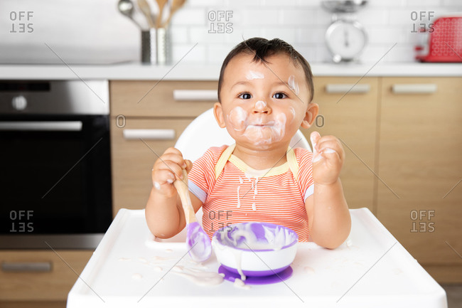 Funny messy baby in high chair eating yogurt with spoon