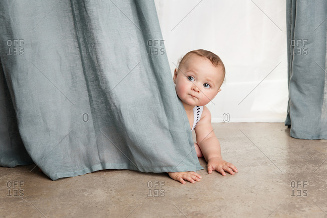 Cute baby crawling on floor peeking behind curtains at home