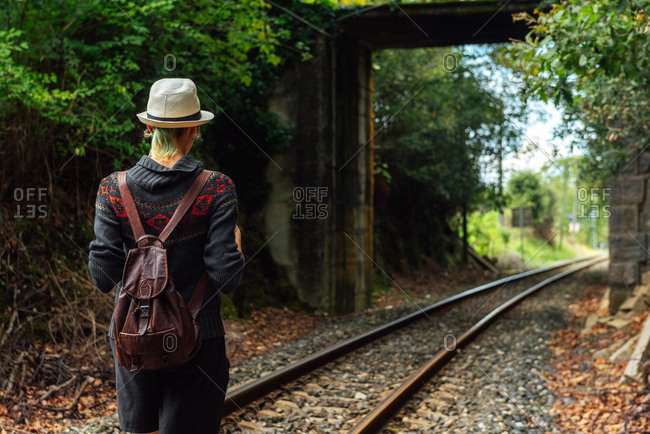 Back view of female traveler with backpack standing on rails in forest during summer adventure