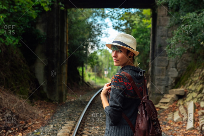 Back view of female traveler with backpack standing on rails in forest and looking at camera over shoulder during summer adventure