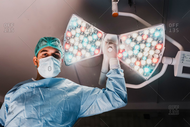 From below of serious male surgeon in protective mask and medical uniform adjusting surgical lamp while preparing for operation in modern operating theater