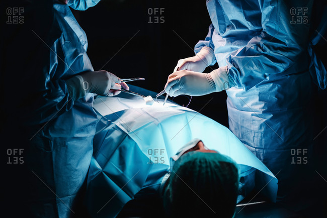 Crop anonymous surgeon and assistant with surgical tools and thread stitching wound of patient during operation in hospital