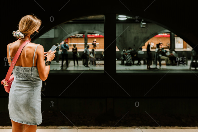 Back view of young female commuter in casual outfit and protective mask for coronavirus prevention checking electronic schedule on smartphone while waiting for train on platform
