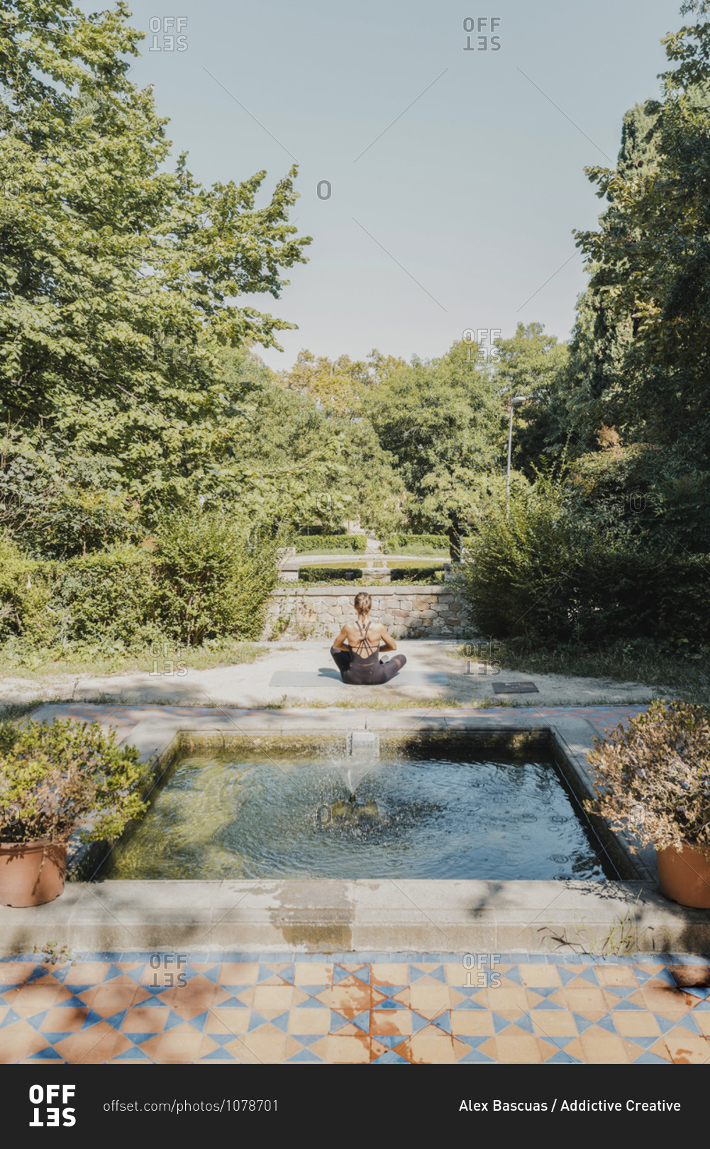 Back view of unrecognizable female sitting in lotus pose and meditating while practicing yoga in green park with water fountain