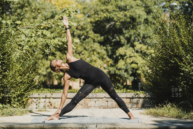 Full body of slim barefoot female in black activewear standing in Triangle position with arm extended while practicing yoga in green park in summer day