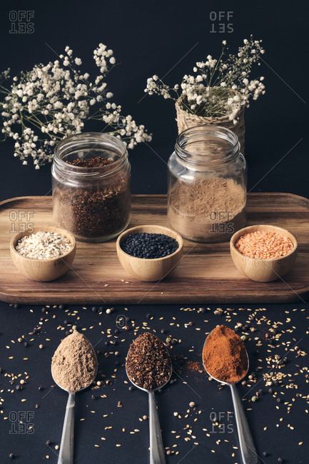 Top view of assorted grains in jars and aromatic spices in spoons arranged on wooden tray on black table