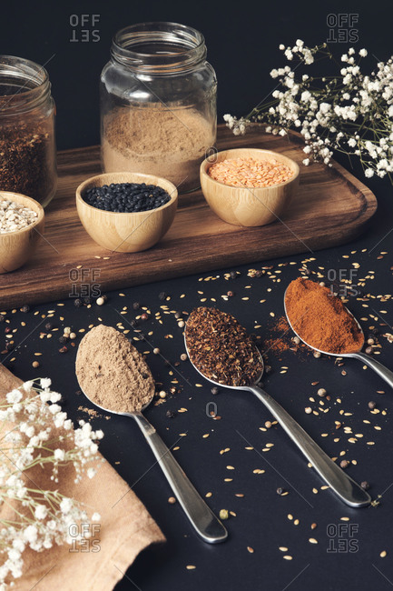 Top view of assorted grains in jars and aromatic spices in spoons arranged on wooden tray on black table