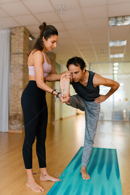 Full body female instructor supporting adult man doing Hasta Padangusthasana pose during yoga lesson in studio