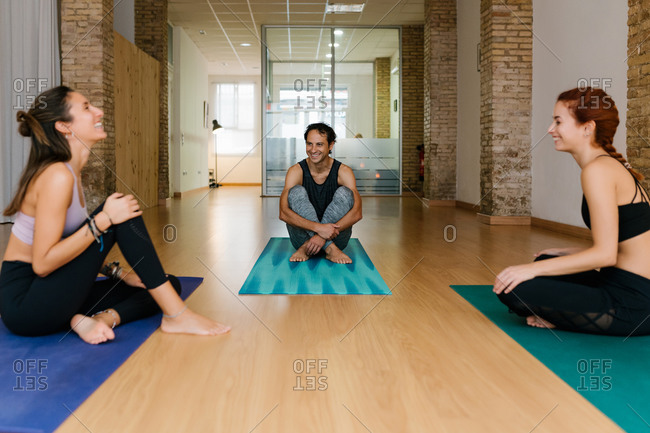 Happy man and women smiling and talking while sitting on mats during break in yoga lesson in studio