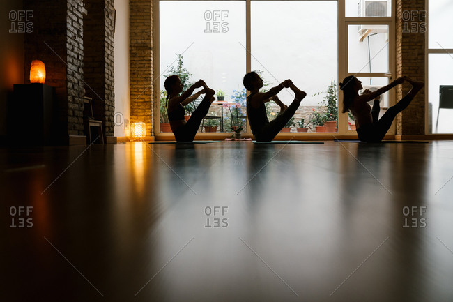 Silhouettes of man and women doing Upavista Konasana pose against window during yoga lesson in dark studio