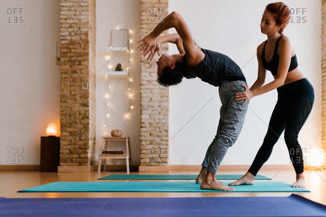 Young female instructor helping adult male to do Wheel pose during yoga lesson in studio