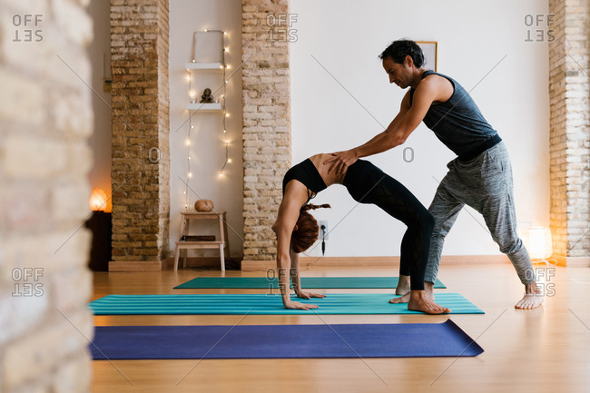 Young male instructor helping adult female to do Wheel pose during yoga lesson in studio