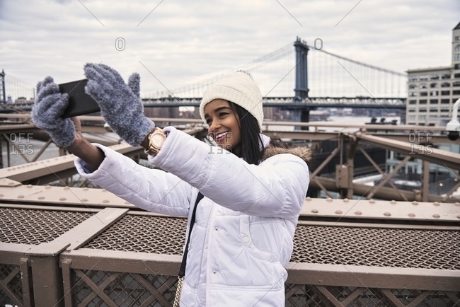 Cheerful African American woman in warm clothes taking photo on selfie camera of smartphone while standing on street of New York
