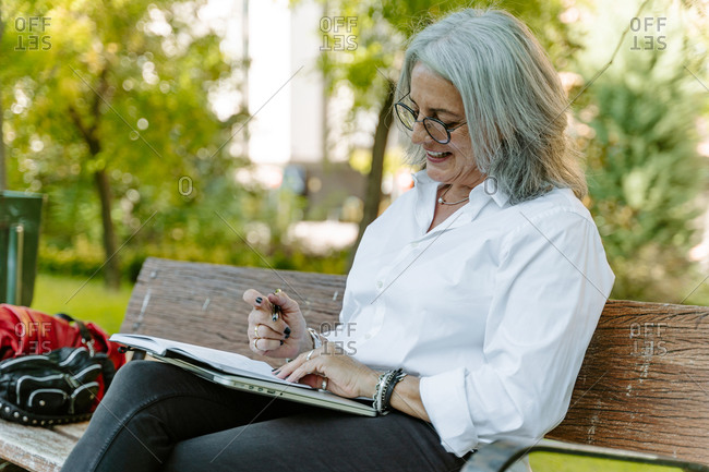Side view of middle aged female entrepreneur in smart casual wear sitting on bench and writing list of daily routine tasks while working remotely in park