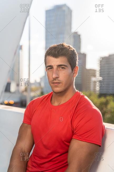 Young muscular male in sportswear resting after intense workout on terrace of modern city building looking at camera