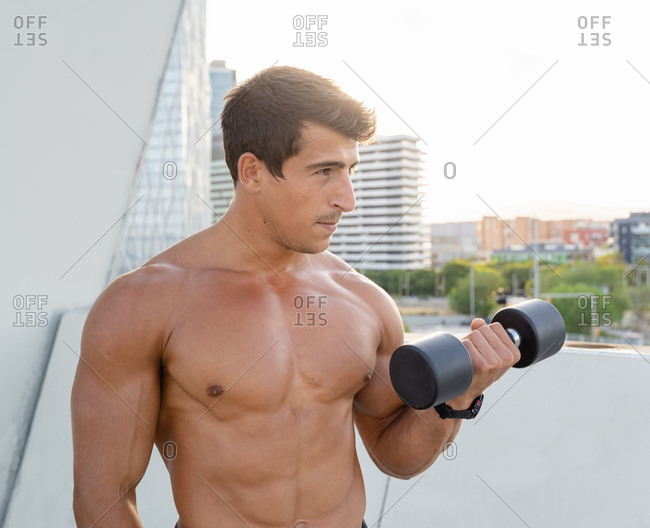 Athletic muscular shirtless guy doing fitness exercise with dumbbell during intense workout in city