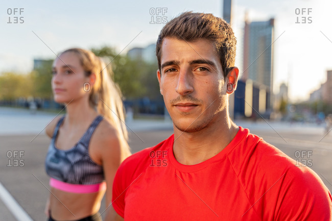 Young trendy male athlete in bright wear looking at camera near fit girlfriend in town in back lit
