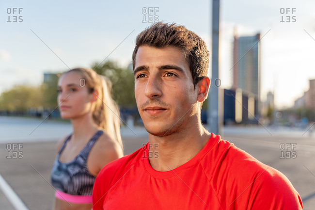 Young trendy male athlete in bright wear looking away near fit girlfriend in town in back lit