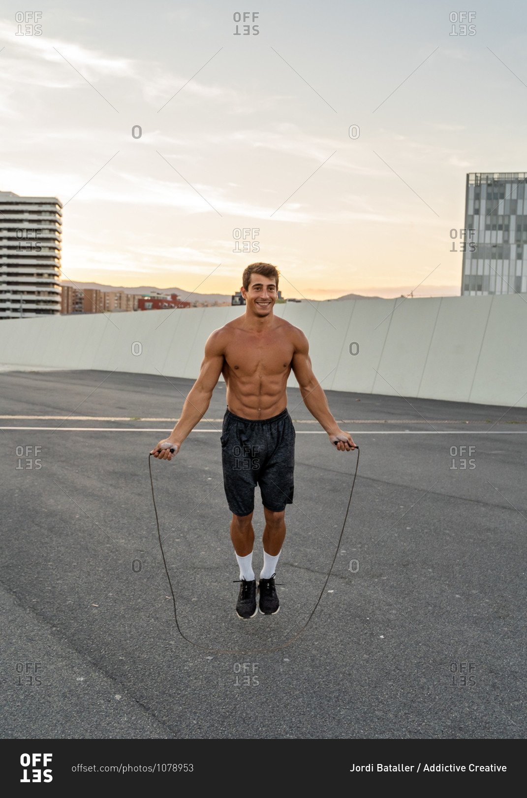 Young smiling muscular male athlete jumping rope on roadway during workout under shiny sky in evening and looking forward