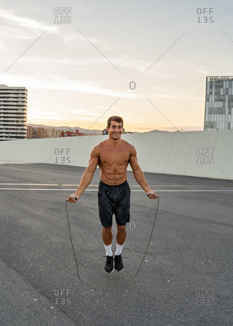 Young smiling muscular male athlete jumping rope on roadway during workout under shiny sky in evening and looking forward