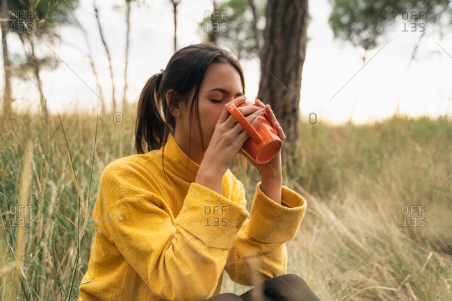 Side view of tranquil female sitting in field in autumn and drinking hot beverage while enjoying weekend