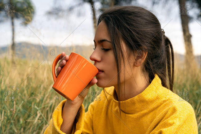 Side view of tranquil female sitting in field in autumn and drinking hot beverage while enjoying weekend