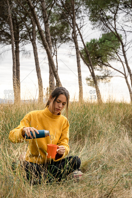 Serene female sitting on dry grass of meadow and pouring hot drink from thermos in cup while enjoying weekend in nature