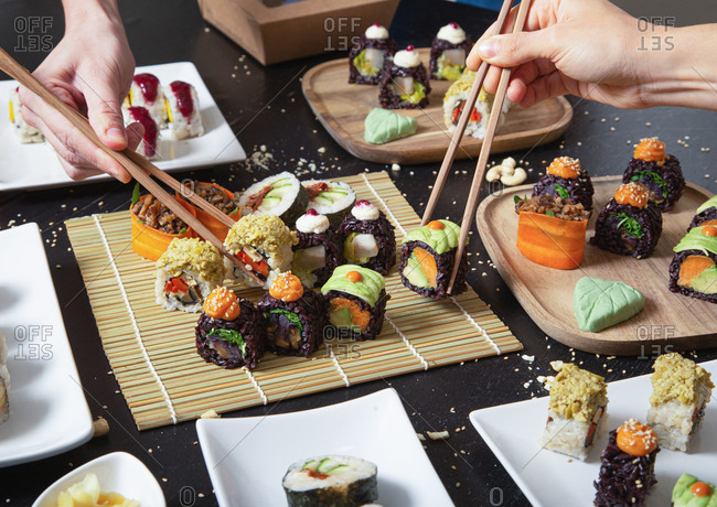 From above of crop unrecognizable people with chopsticks taking assorted veggie sushi from bamboo mat