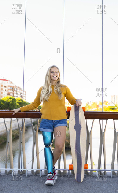 Carefree female skater in summer clothes and with bionic leg prosthesis standing on stone border in city and looking away