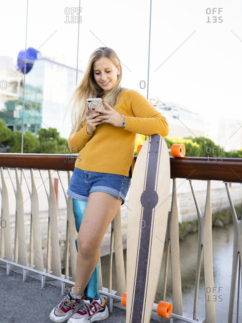 Delighted female skater with bionic leg prosthesis standing on bridge with longboard and reading messages on social media on smartphone