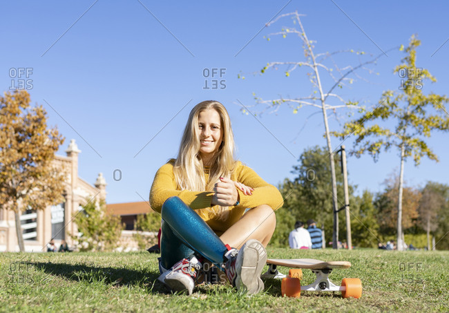 Joyful woman with leg artificial limb sitting on grass in park with longboard and relaxing on sunny day