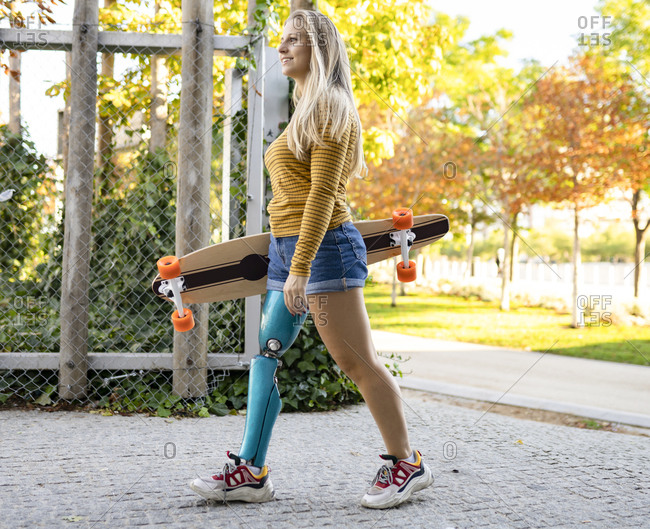 Positive female skater with bionic leg prosthesis walking with longboard in city and looking away