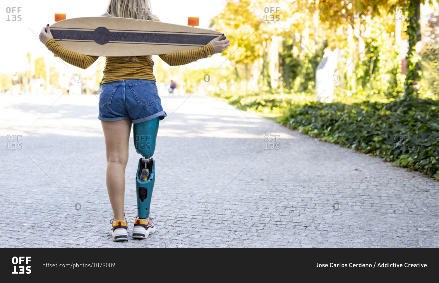 Back view of crop anonymous female skater with bionic prosthesis standing in city with longboard and enjoying summer