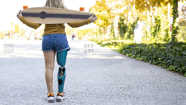 Back view of crop anonymous female skater with bionic prosthesis standing in city with longboard and enjoying summer