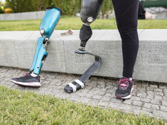 Unrecognizable crop female runner changing leg prosthesis for training while sitting on stone border in urban park