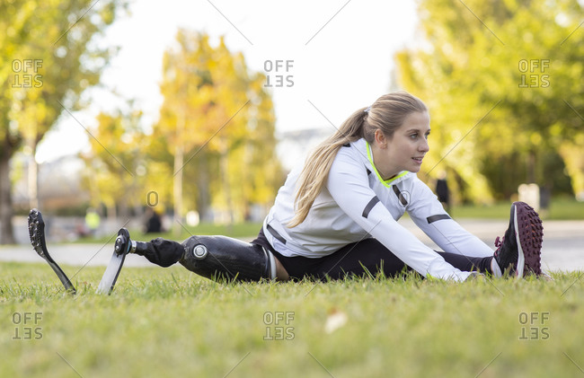 Professional female sportswoman with artificial limb sitting on grass in park and stretching legs while doing forward bend during workout