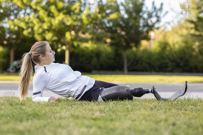 Side view of female Paralympic runner with artificial limb stretching legs during training in park while warming up and looking away