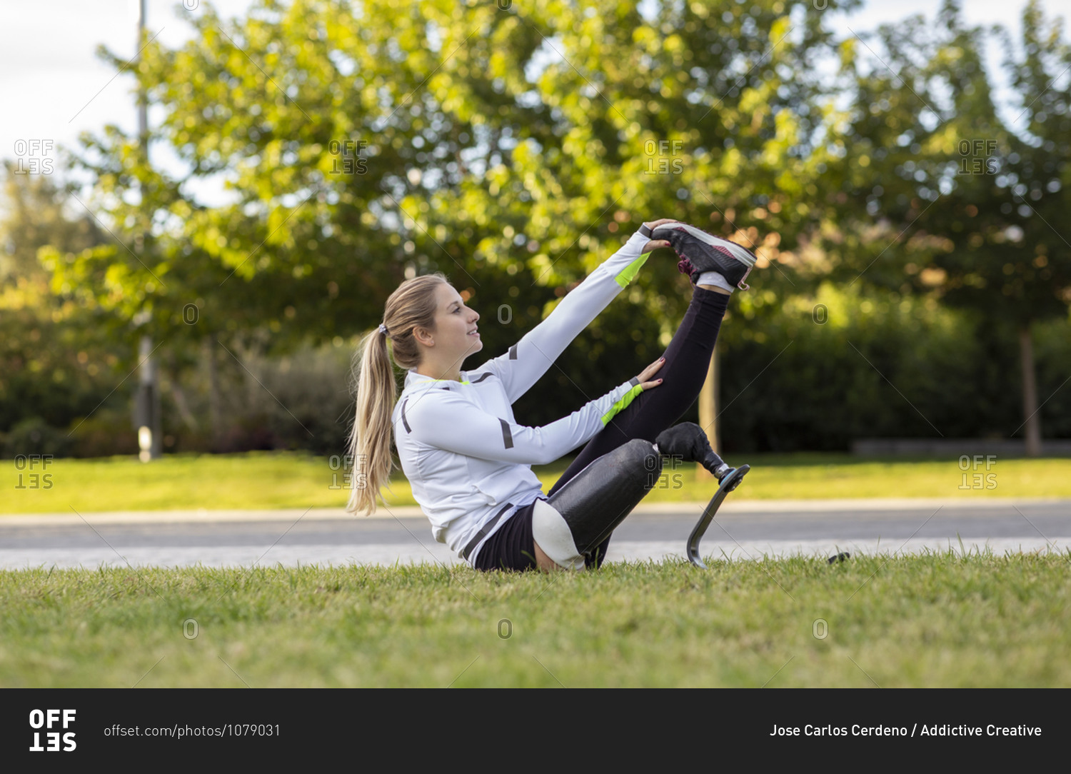 Side view of professional female runner with bionic prosthesis stretching legs during workout while sitting on grass