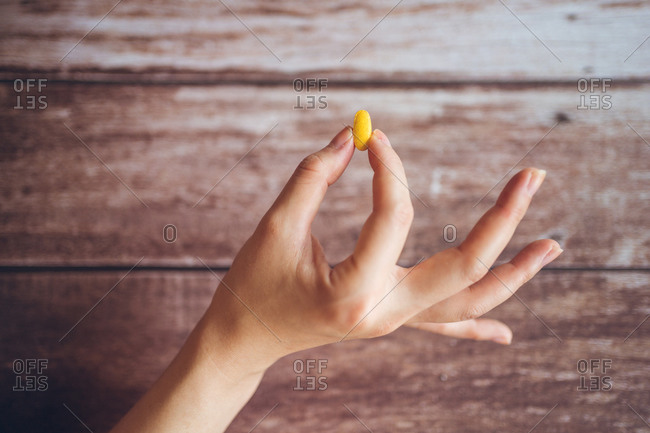 Crop hand of anonymous female demonstrating bright yellow supplement pill for healthy eating and lifestyle concept against shabby wooden background