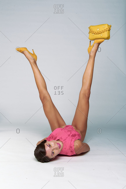 Full body of cheerful young fashionable female model in pink wear and yellow high heels lying on floor with small yellow purse on raised leg in white studio