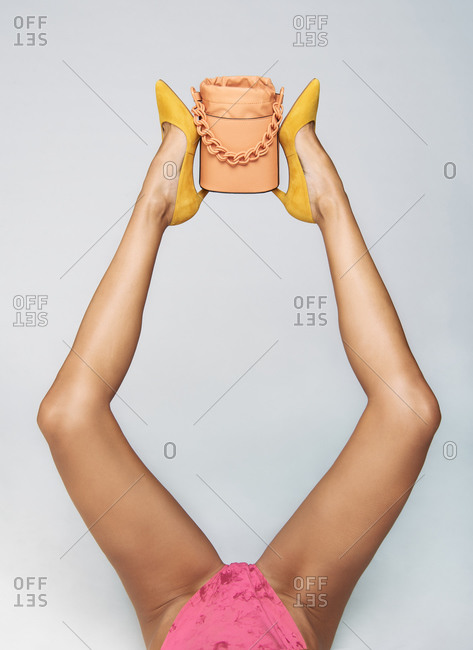 Cropped unrecognizable fashionable female model in pink wear and yellow high heels lying on floor with small yellow purse on raised leg in white studio