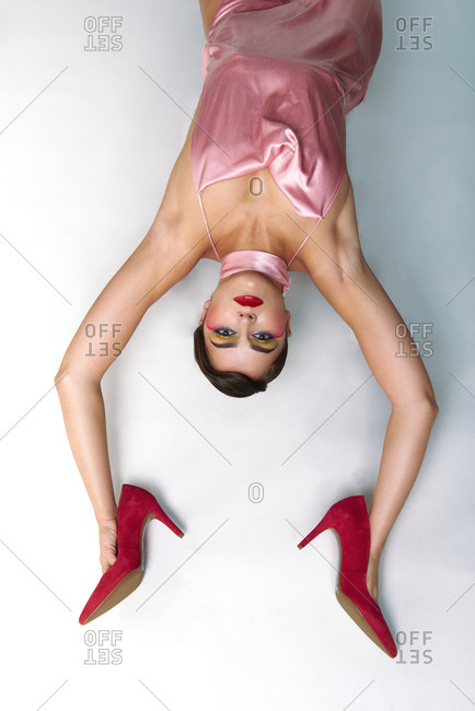 From above of fashionable female model with bright makeup wearing pink dress holding stylish red high heeled shoes in hands while lying on white floor