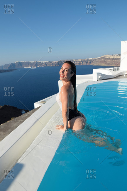 Side view of delighted female tourist in swimsuit and with wet hair leaning on poolside of swimming pool and enjoying vacation on Santorini while looking at camera