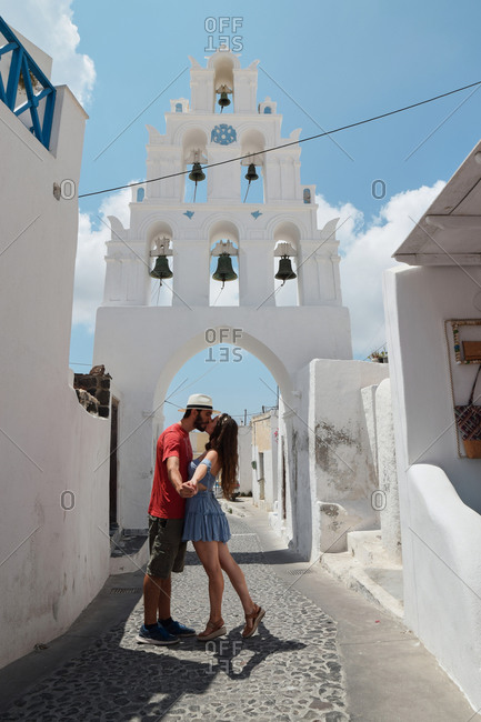 Side view of couple of tourists standing on old narrow street near church and tenderly kissing during summer vacation on Santorini