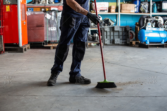 Low angle of man in uniform sweeping floor with broom while working in garage