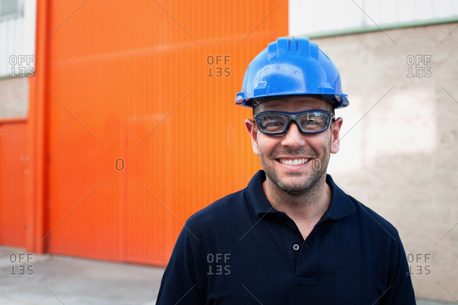 Positive confident adult male workman in blue protective hardhat and eyeglasses smiling friendly while standing against bright orange wall of workshop